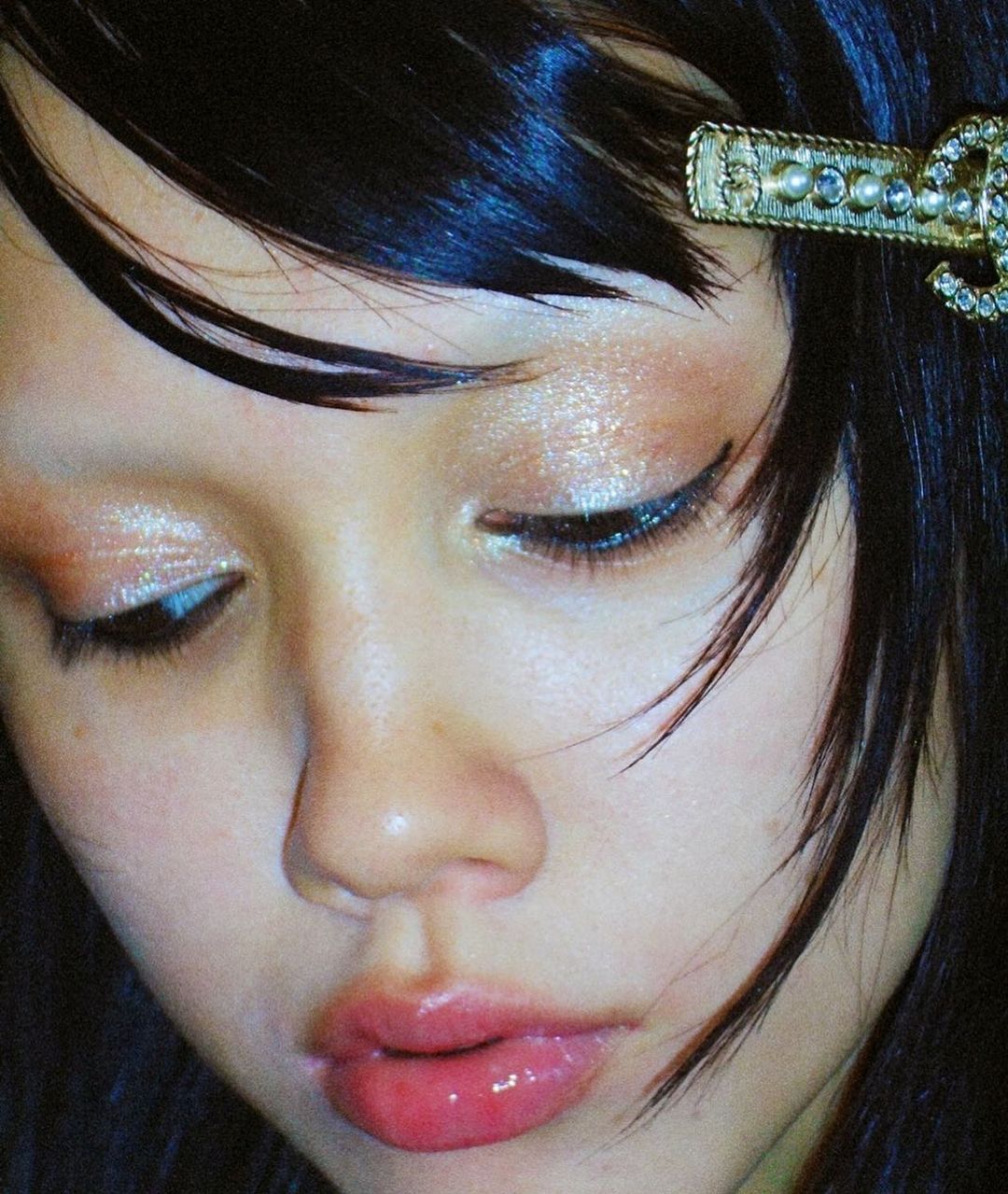 Close-up beauty shot of an adult woman with dark hair styled with a decorative gold hair clip, wearing shimmering eye makeup and glossy lipstick.