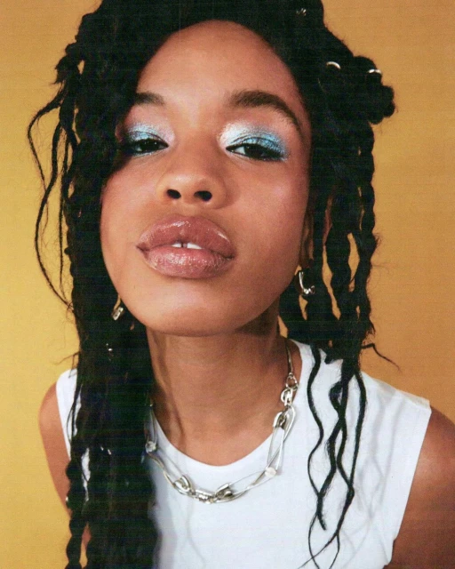 Close-up portrait of a Black female model with locs, glossy lips, and blue eyeshadow, wearing a white top and silver chain against a yellow backdrop.