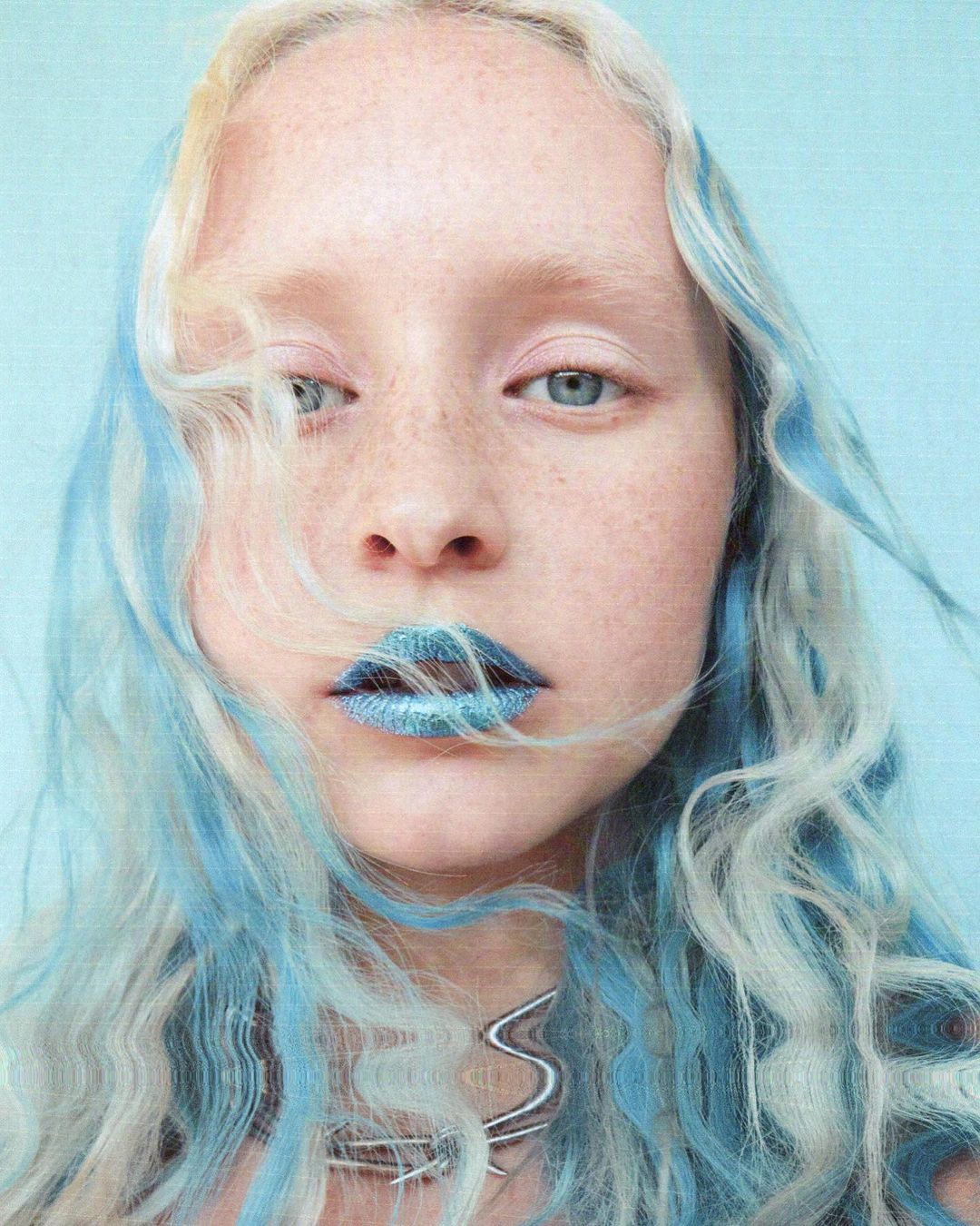 Close-up of a young adult female with white-blonde hair and light blue highlights, wearing sparkling blue lipstick and a silver necklace.