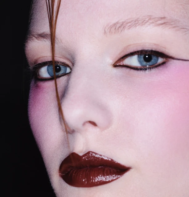 Close-up portrait of a woman with blue eyes and glossy dark red lips, accentuated by pink blush and precise black eyeliner.