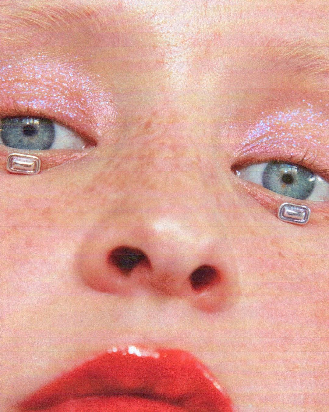 Close-up portrait of a fair-skinned woman with glossy red lips, glittery pink eyeshadow, and small silver rectangles under her eyes.