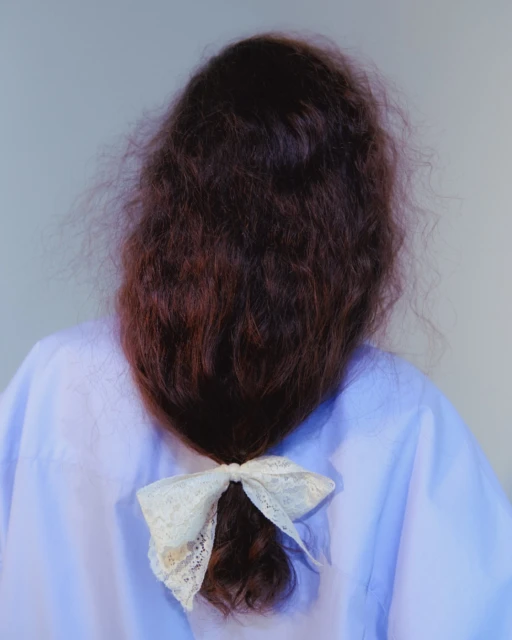 Back view of a woman with wavy auburn hair tied in a low ponytail with a white lace bow, wearing a light purple button-down shirt.