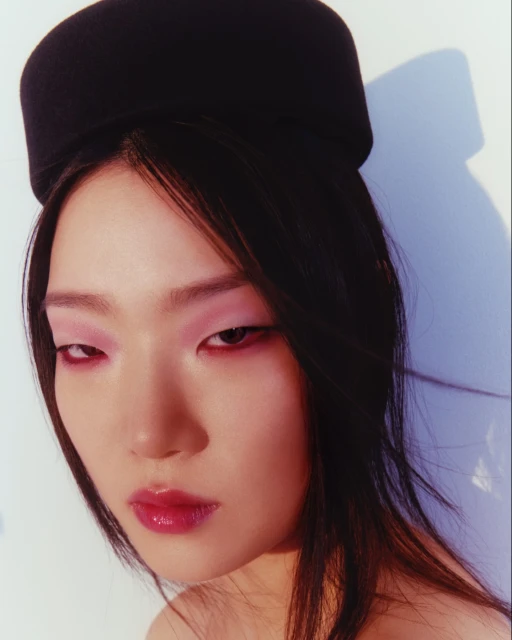 Close-up of an Asian woman with pink eye shadow and red lipstick wearing a black hat against a white and light blue background.
