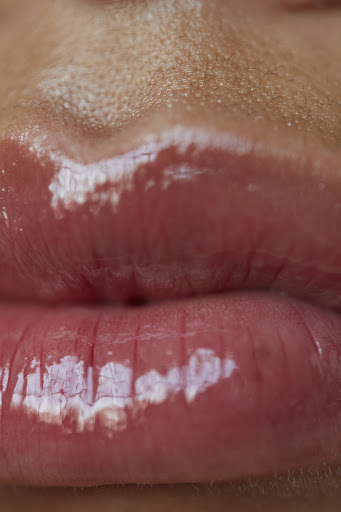 Close-up of a person's mouth with glossy pink lipstick. The skin is visible above the lips, and light reflects off the glossy surface.
