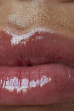 Close-up of a person's mouth with glossy pink lipstick. The skin is visible above the lips, and light reflects off the glossy surface.
