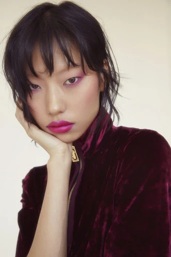 Portrait of an East Asian woman with dark hair, vibrant pink makeup, and a dark velvet jacket against a cream-colored backdrop in a studio setting.