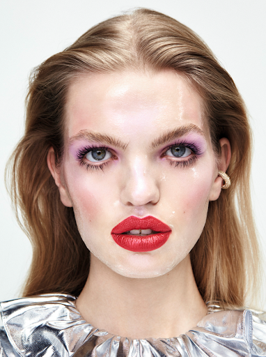 Close-up portrait of a woman with light brown hair, dramatic red lipstick, and iridescent eyeshadow on a white background.