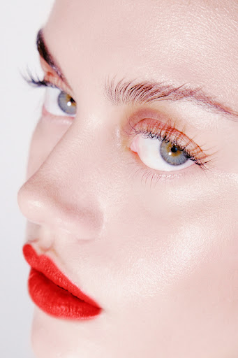 Close-up portrait of a light-skinned woman's face featuring bright red lipstick, full lashes, well-groomed eyebrows, and grey-green eyes.