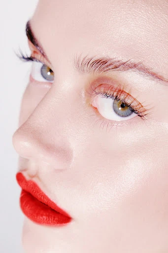 Close-up portrait of a light-skinned woman's face featuring bright red lipstick, full lashes, well-groomed eyebrows, and grey-green eyes.