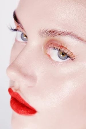 Close-up portrait of a light-skinned woman's face featuring bright red lipstick, full lashes, well-groomed eyebrows, and grey-green eyes.