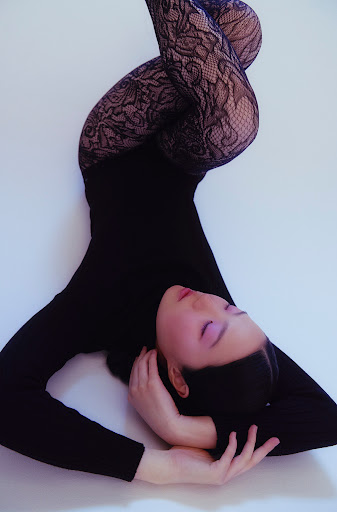 Woman in black bodysuit and lace leggings poses with arms crossed behind her head against a white backdrop in a studio shot.