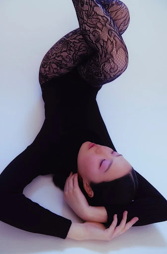 Woman in black bodysuit and lace leggings poses with arms crossed behind her head against a white backdrop in a studio shot.