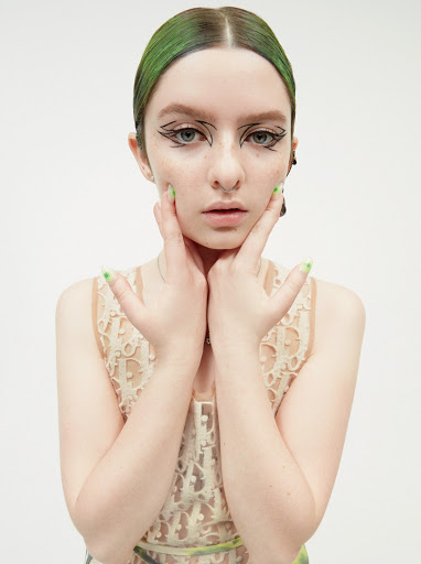 Headshot of a young woman with green hair and graphic eyeliner posing with her hands framing her face in a bright studio setting.