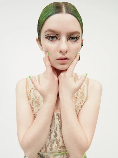 Headshot of a young woman with green hair and graphic eyeliner posing with her hands framing her face in a bright studio setting.