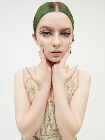 Headshot of a young woman with green hair and graphic eyeliner posing with her hands framing her face in a bright studio setting.