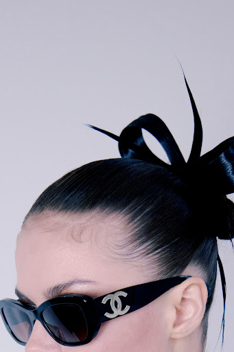 Close-up fashion shot: woman wears black sunglasses with silver logo, black hair pulled back with dark bow accent, and a light grey backdrop.