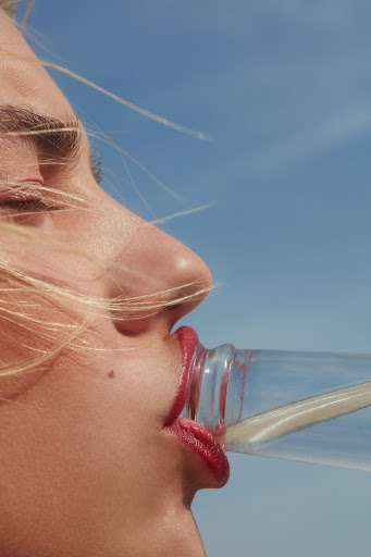 Close-up of a woman drinking from a clear glass bottle, her eyes closed, fair skin, with a clear blue sky background.