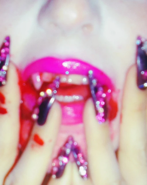 Close-up of woman's face with glossy pink lips, vampire fangs, and fingers with long, decorated nails near her mouth in an indoor setting.