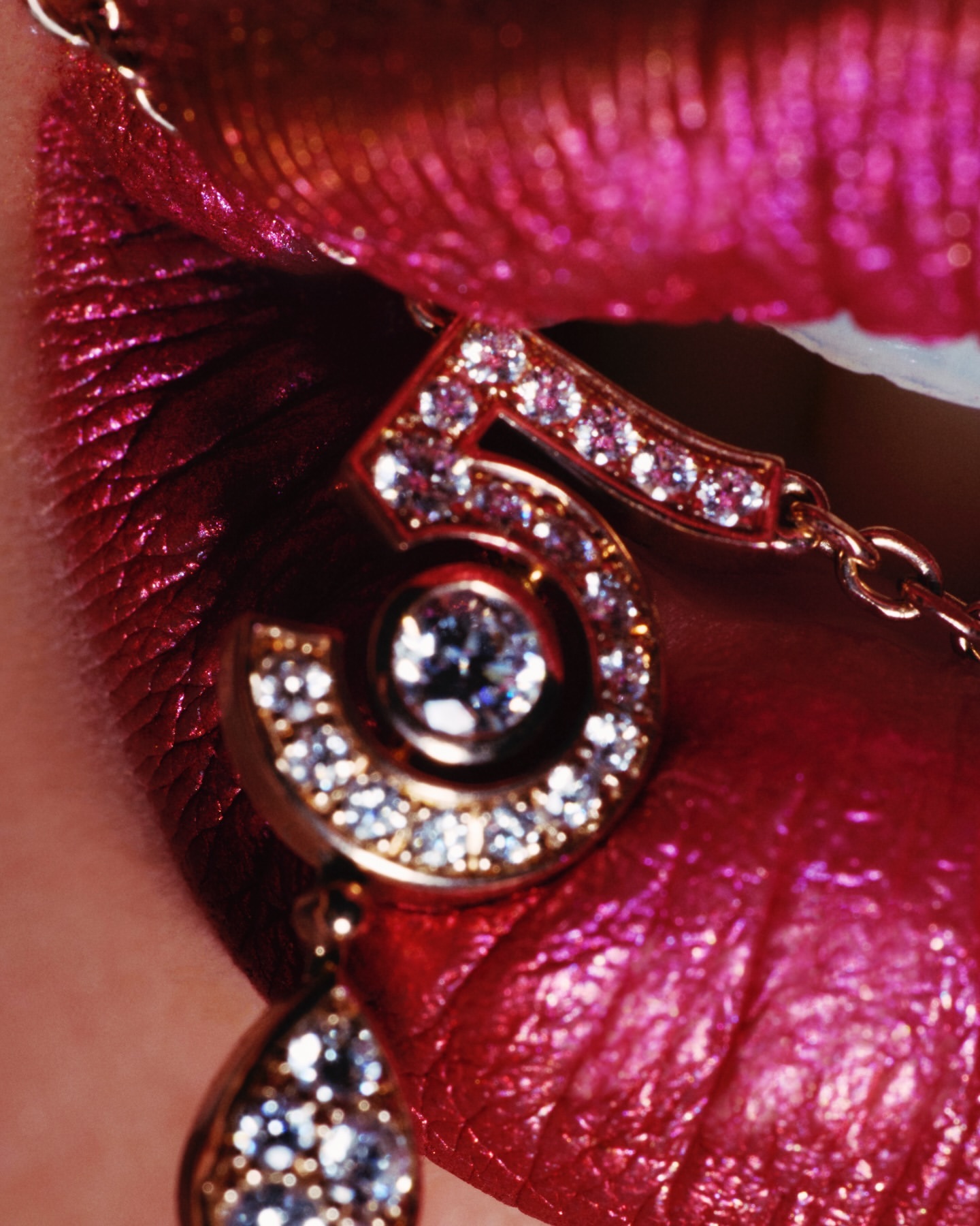 Close-up shot of a woman's shiny pink lips with gold chain jewelry that includes a glittering number 5 pendant.