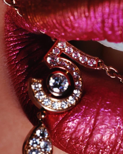 Close-up shot of a woman's shiny pink lips with gold chain jewelry that includes a glittering number 5 pendant.