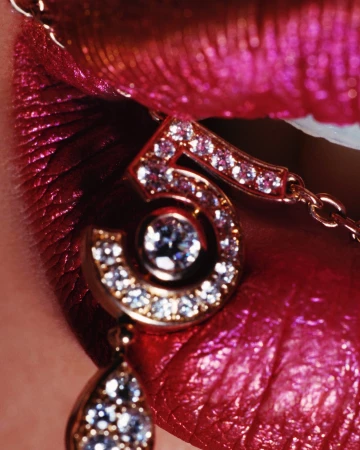 Close-up shot of a woman's shiny pink lips with gold chain jewelry that includes a glittering number 5 pendant.