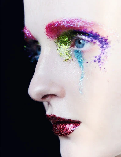 Close-up of a woman's face featuring colorful glitter makeup on the eyelids and lips, set against a dark background.