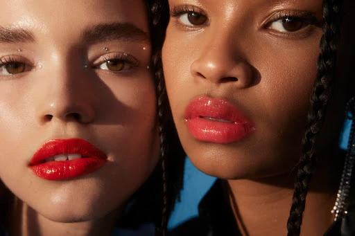 Close-up of two women showcasing red lip gloss. Beauty shot with studio lighting highlighting glossy lips, clear skin, and cosmetic details.