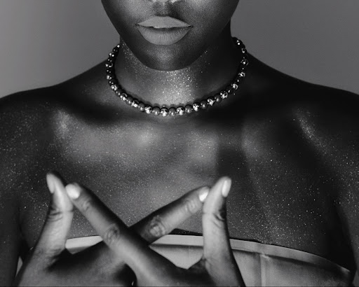 Black and white studio portrait of African American woman with a beaded necklace and hands crossed, showing shoulders and upper chest.