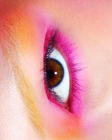 Extreme close-up of a brown eye featuring vibrant pink eyeshadow and long dark eyelashes with warm orange brow tones.