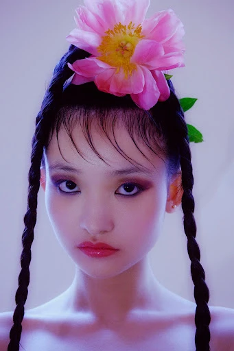 Portrait of a woman with braided hair and a pink peony flower on her head, featuring makeup, glossy lips, and an even skin tone.