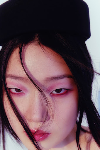 Close-up portrait of a woman wearing dramatic pink eyeshadow and a black structured hat against a plain background