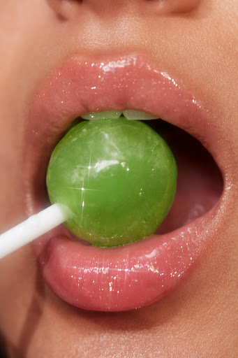 Close-up of a person with pink lipstick holding a vibrant green lollipop in their mouth, illuminated by a bright and shiny light.