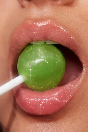 Close-up of a person with pink lipstick holding a vibrant green lollipop in their mouth, illuminated by a bright and shiny light.