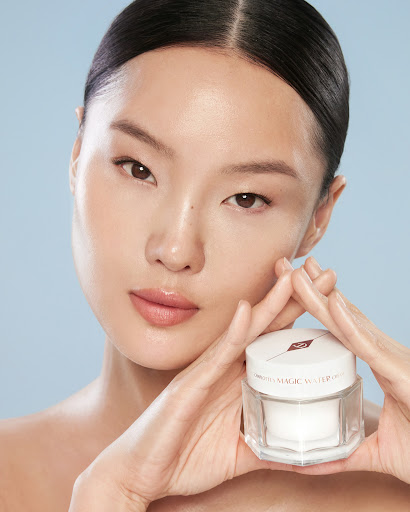Close-up of an Asian woman with smooth skin holding a jar of Charlotte Tilbury Magic Water Cream against a light blue backdrop.