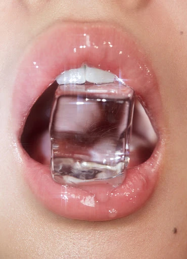 Close-up shot of an adult's mouth holding a transparent ice cube between their teeth and lips, set against a neutral background.