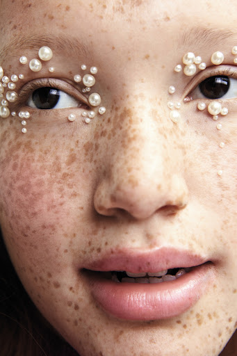 Close-up of a young person's face adorned with small pearls around the eyes, highlighting their freckled skin and natural beauty.