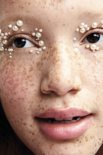 Close-up of a young person's face adorned with small pearls around the eyes, highlighting their freckled skin and natural beauty.