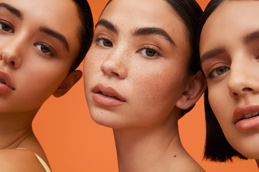 Close-up of three diverse young women with flawless skin posing in front of an orange backdrop, emphasizing beauty and skincare.