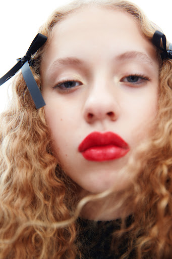 Close-up portrait of a young girl with curly blonde hair and red lipstick, adorned with black bows in her hair, against a white background.