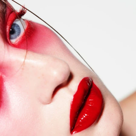 Close-up beauty portrait of a woman with vibrant red eyeshadow, bright red lipstick, and striking blue eyes against a white background.