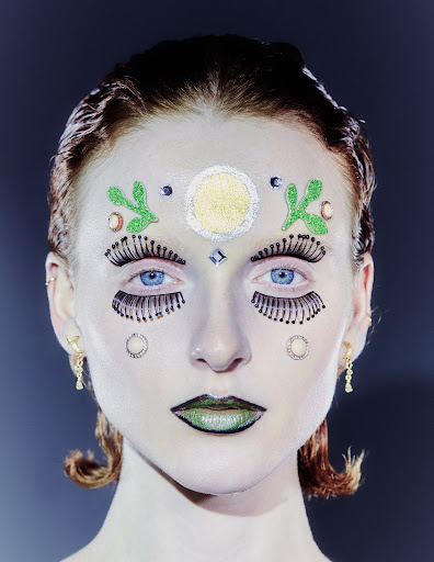 Close-up of a person with face glitter, false eyelashes, and creative makeup. The individual has light skin, blue eyes, and auburn hair.