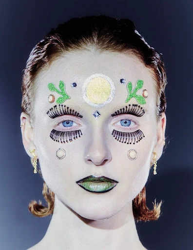 Close-up of a person with face glitter, false eyelashes, and creative makeup. The individual has light skin, blue eyes, and auburn hair.