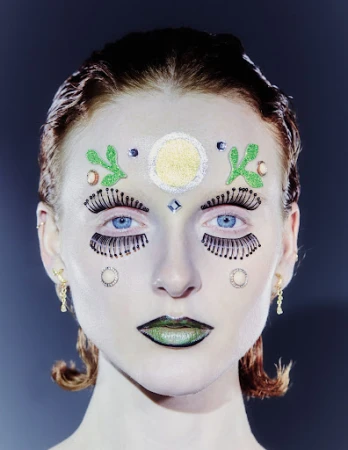 Close-up of a person with face glitter, false eyelashes, and creative makeup. The individual has light skin, blue eyes, and auburn hair.