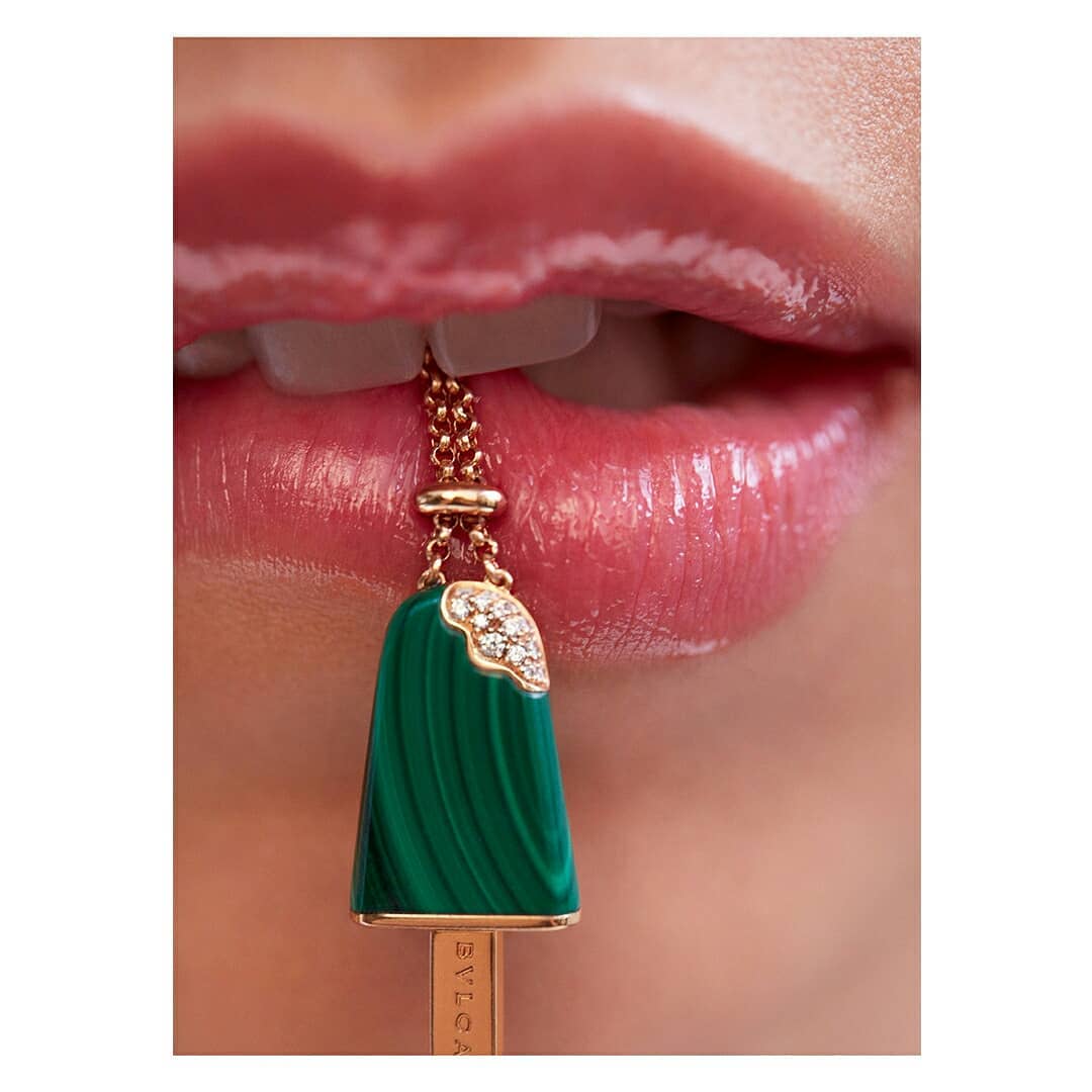 Close-up of a woman's glossy pink lips and teeth with a green malachite and diamond pendant necklace between the teeth.
