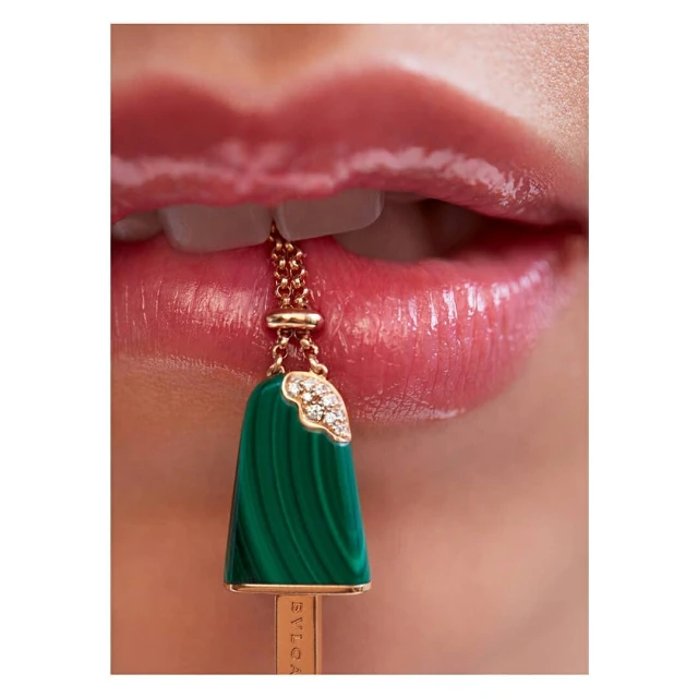 Close-up of a woman's glossy pink lips and teeth with a green malachite and diamond pendant necklace between the teeth.