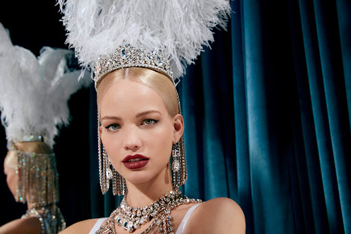 Close-up of a blonde woman wearing a crystal tiara, feathered headpiece, and heavy rhinestone jewelry against a dark teal curtain.