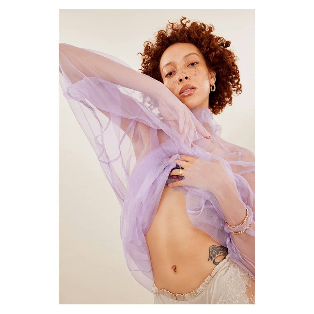 A young adult woman poses in a studio wearing a sheer lavender top, lace shorts, and delicate jewelry against a neutral background.