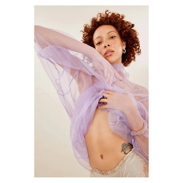 A young adult woman poses in a studio wearing a sheer lavender top, lace shorts, and delicate jewelry against a neutral background.