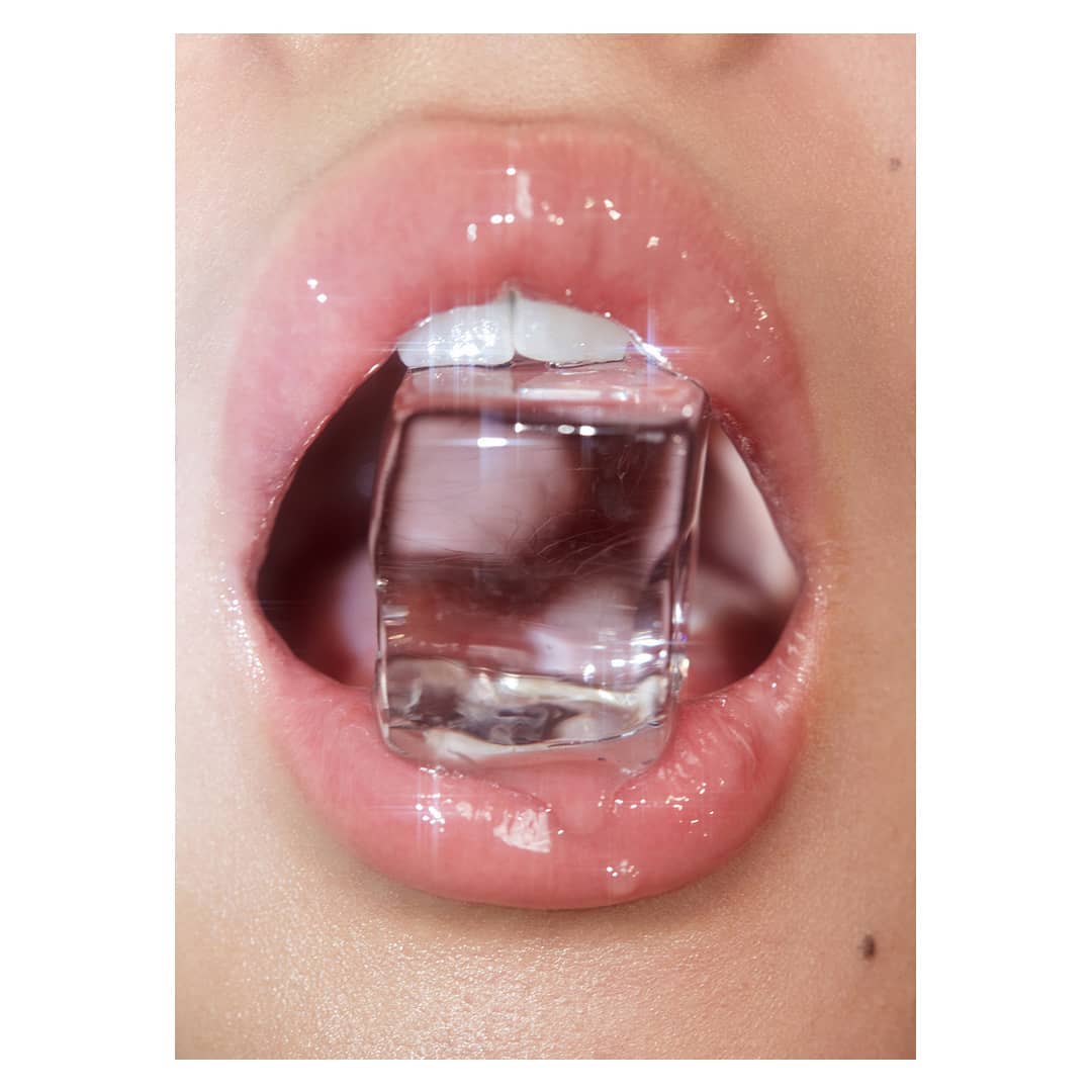 Close-up of a person's mouth with glossy lips and an ice cube held between the teeth, creating a refreshing and eye-catching visual.