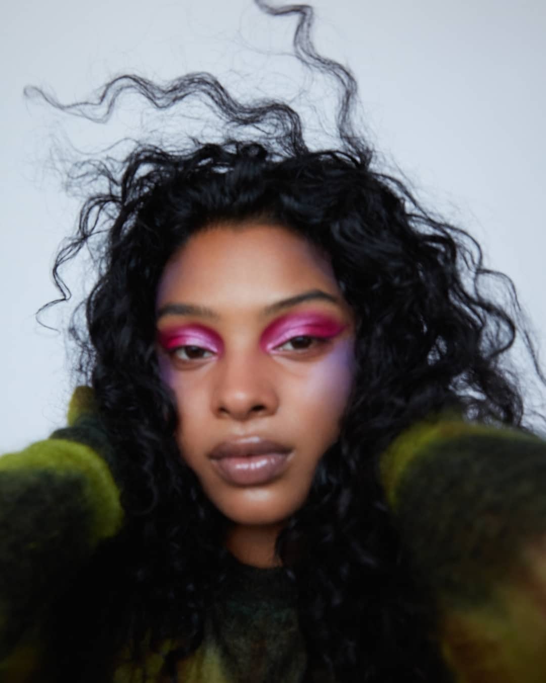 Close-up portrait of a woman with long dark curly hair wearing vibrant pink eyeshadow against a white background by Anairam